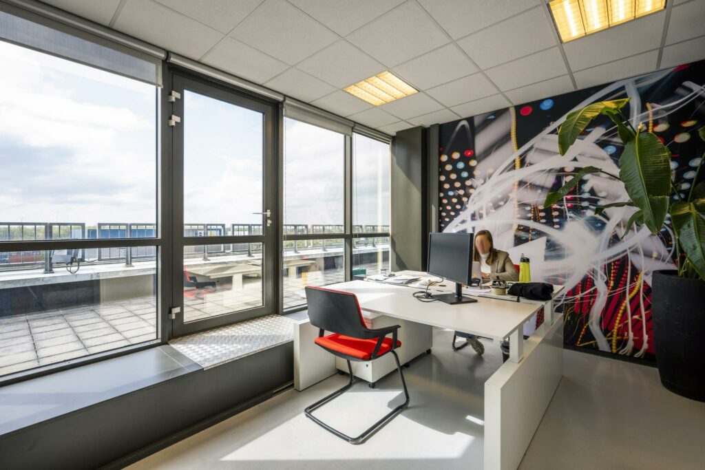 Modern kantoor met grote ramen, een kleurrijke wanddecoratie en een persoon werkend aan een bureau aan de Anthony Fokkerweg.